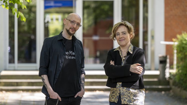 Eine Frau mit verschränkten Armen, ein Mann mit Händen in den Taschen, stehen vor einem Gebäude.