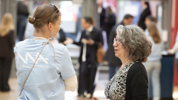 A person wearing a light blue Northwest Alliance T-shirt can be seen from behind, talking to a woman wearing glasses and with medium-length grey hair. 
