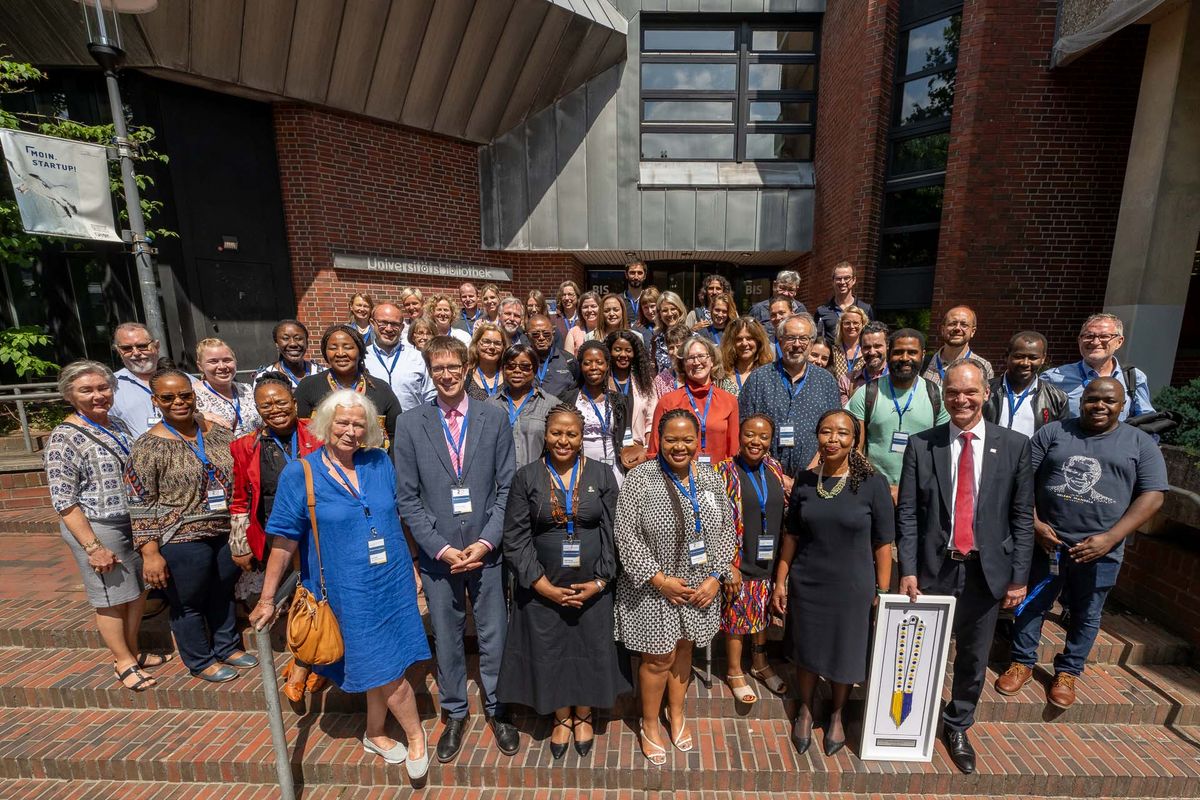 Das Bild zeigt eine große Gruppe von Menschen, die vor dem Eingang der Universitätsbibliothek der Universität Oldenburg posieren. Es handelt sich um eine Gemeinschaft von Dozierenden, Mitarbeitenden und dem Präsidium der Universität Oldenburg und der Nelson Mandela University, die anlässlich des 25-jährigen Kooperationsjubiläums zusammengekommen sind. Die Gruppe steht auf einer Treppe vor einem modernen Gebäude mit Glasfassade und Bäumen im Hintergrund. Viele tragen Namensschilder und formelle Kleidung. Das Bild vermittelt einen Eindruck von internationaler Zusammenarbeit und festlicher Stimmung.