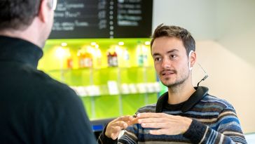 Ein junger Mann erklärt einem anderen Mann etwas, an seinem Ohr ist ein C-förmiges EEG-Messgerät befestigt.