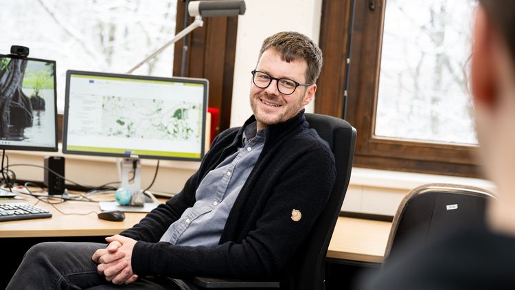 Das Bild zeigt Thorsten Balke in seinem Büro. Er sitzt auf einem Schreibtischstuhl und lächelt den Interviewer an. Hinter ihm befinden sich zwei Bildschirme, einer davon zeigt die Verteilung von Grünflächen in der Stadt Oldenburg.