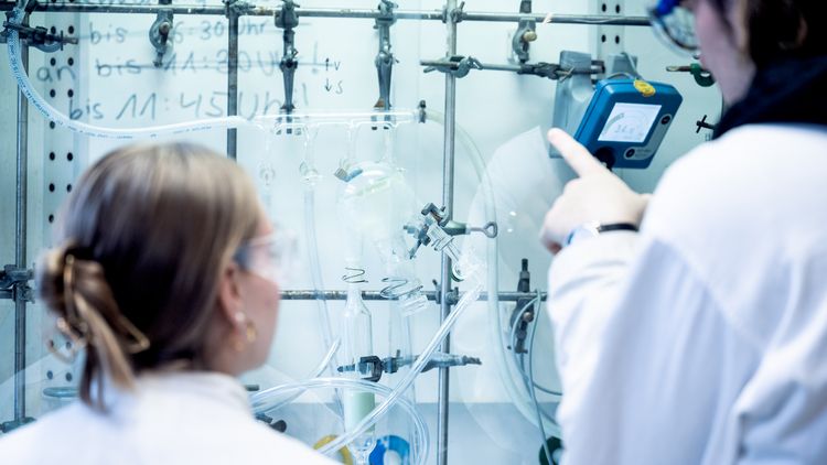 Zwei Personen stehen vor einem Aufbau mit verschiedenen chemischen Geröten, die durch Schläuche verbunden sind.