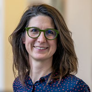A lady with brown, shoulder-length hair and a blue blouse with red dots smiles at the camera with her mouth open. She wears striking green glasses.
