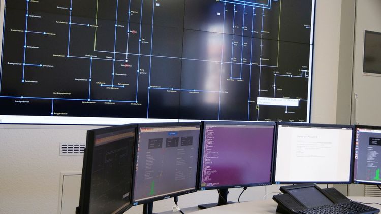 A large screen with a circuit diagram on the wall, below which several large computer screens are lined up next to each other, similar to a control centre