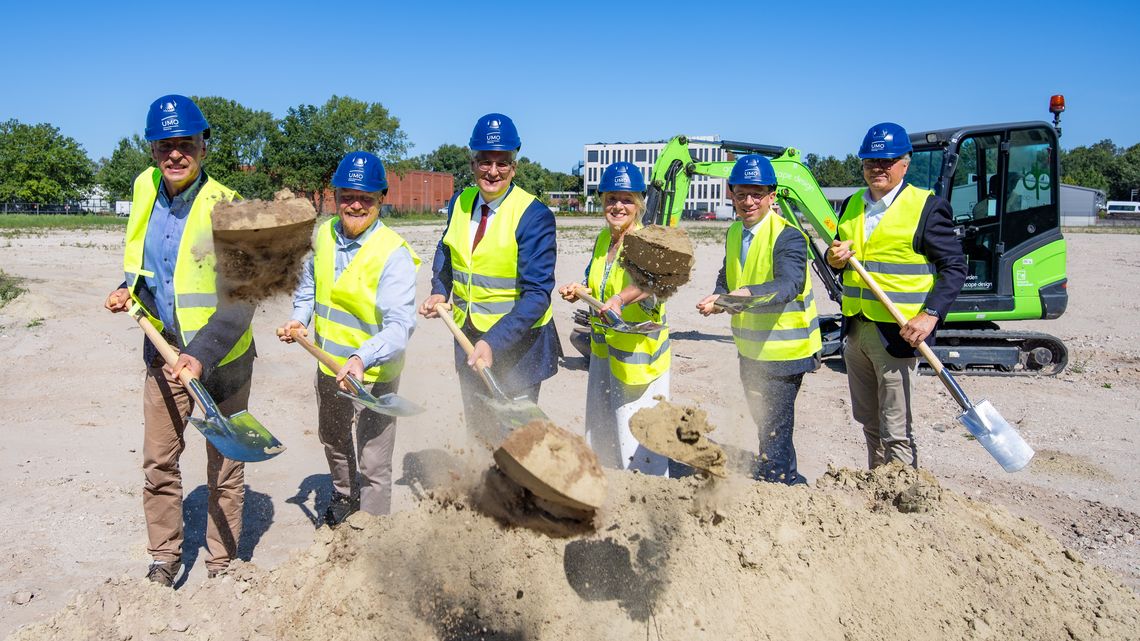 The people pictured are wearing yellow high-visibility vests and blue helmets bearing the UMO logo, and are shovelling sand towards the camera.