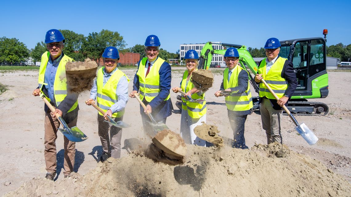 Die genannten Personen tragen gelbe Warnwesten, blaue Helme mit UMO-Logo und schippen Sand Richtung Kamera.