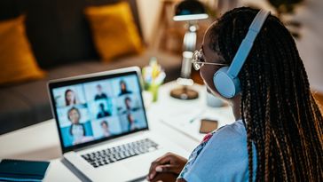 Young woman with rasta braids and headphones sits in front of a laptop. Participants in a video conference can be seen on the screen.