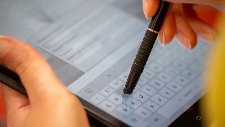 Großaufnahme eines Tabletts. Zwei Hände sind sichtbar, die linke hält das Tablett, die recht hält einen Spezialstift, mit der auf dem Bildschirm etwas eingegeben wird. 