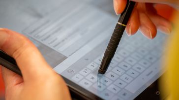 Großaufnahme eines Tabletts. Zwei Hände sind sichtbar, die linke hält das Tablett, die recht hält einen Spezialstift, mit der auf dem Bildschirm etwas eingegeben wird. 