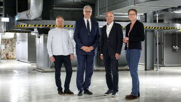 Vier Personen in förmlicher Kleidung stehen im Technikraum vor großen Luftkanälen und schauen in die Kamera.