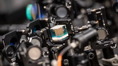 Close-up of the experimental setup, in the centre a transparent glass cube, surrounded by various small devices that redirect or reflect laser beams.