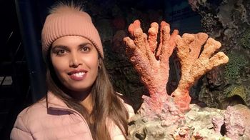 Photo of Rimaa Beesoo taken at the aquarium in Genoa, Italy. Beesoo is wearing a pink cap and a pink jacket. She is standing next to a model of a coral reef.