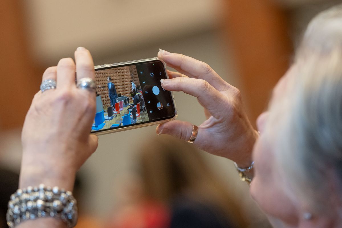 Das Bild zeigt die Hände einer Person, die ein Smartphone benutzt, um ein Foto zu machen. Auf dem Bildschirm des Smartphones ist ein Bild von einem Innenraum mit Menschen zu sehen. Die Person trägt mehrere Ringe an ihren Fingern und mehrere Armbänder. Das Bild vermittelt den Eindruck, dass jemand einen Moment des Symposiums festhält. Der Fokus liegt auf dem Smartphone und dem Prozess des Fotografierens.