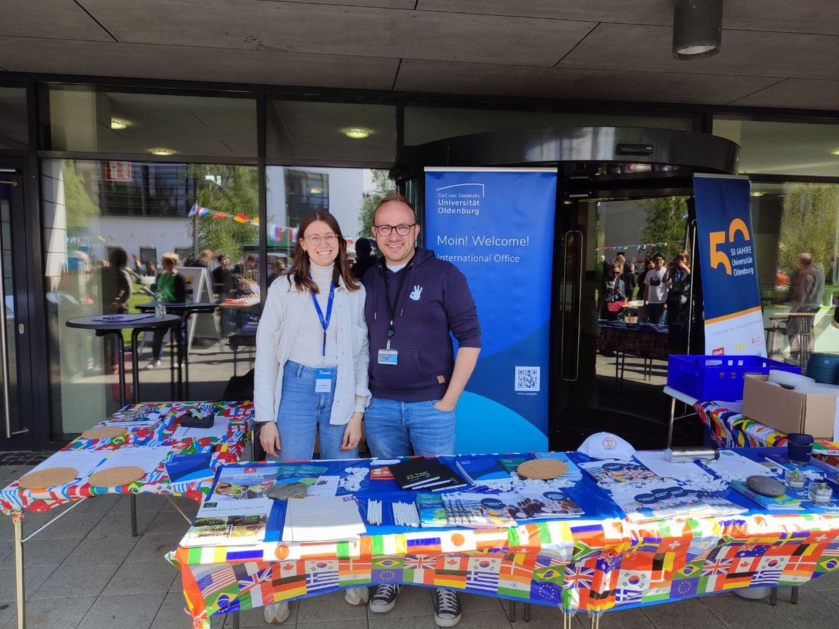 The photo shows two smiling people, a woman and a man, standing in front of a stall. Behind them is a banner with the logo of the University of Oldenburg and the text 'Moin! Welcome! International Office'. The stall is covered with information materials and flyers, including a tablecloth with flags of various countries. In the background, an entrance area and further people can be seen, who are reflected in the glass. The image conveys the impression of a friendly welcome and an information stall of the International Office of the university.