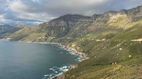 Eine weite Landschaft mit Küstenlinie, Bergen und blauem Meer. Im Hintergrund bewölkte Himmel und grüne Vegetation.