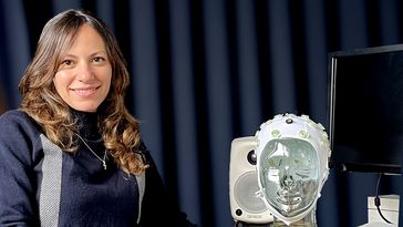 A woman with long hair sits at a table and looks into the camera, next to her a glass head with an EEG cap, a screen and a loudspeaker can be seen. 
