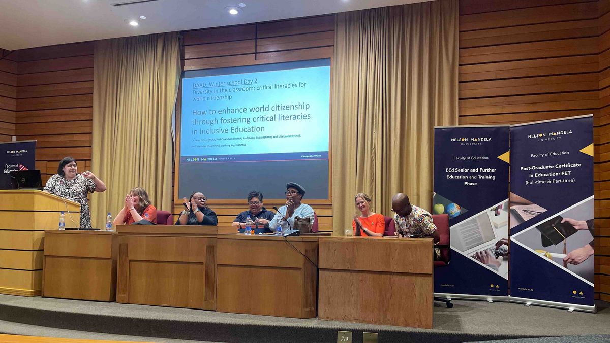 A podium discussion or presentation in a lecture hall. Several people sit behind a podium, one person speaks into a microphone.