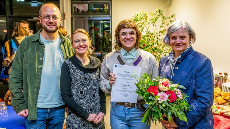 Vier Personen stehen in einem Raum, eine Person hält eine Urkunde und einen Blumenstrauß. Im Hintergrund eine Pflanze und eine geschäftige Atmosphäre.