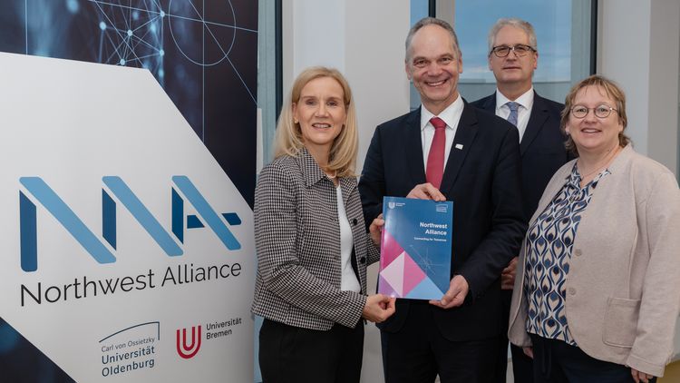 The four people are standing in front of a roll-up banner for the Northwest Alliance, holding the application in their hands.