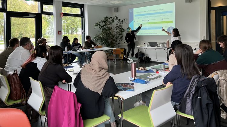 A snapshot of the workshop: participants are seated at U-shaped tables and are looking at a presentation. The two speakers are pointing at something on the projected image.