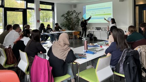 A snapshot of the workshop: participants are seated at U-shaped tables and are looking at a presentation. The two speakers are pointing at something on the projected image.
