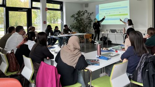 Aufnahme der Workshop-Situation: Teilnehmende sitzen an U-förmig aufgebauten Tischen und sehen auf eine Präsentation. Die beiden Vortragenden zeigen etwas auf dem projizierten Bild.