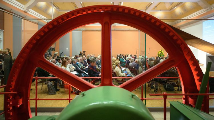 Blick durch ein großes, rotes Zahnrad auf den Saal mit den Zuschauern. 