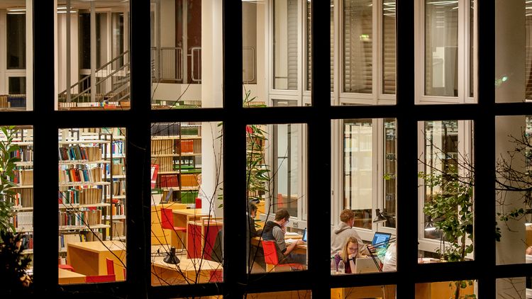 Das Bild zeigt die Bibliothek abends und von außen. In einem Raum kann man Menschen erkennen, die an Schreibtischen arbeiten. im Hintergrund stehen lange Regalreihen mit Büchern.