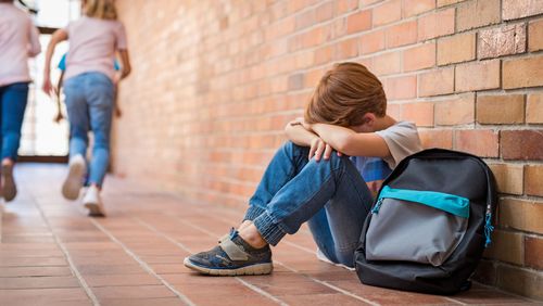 Junge im Grundschulalter sitzt auf einem Schulflur und verbirgt das Gesicht zwischen den Armen,  andere Kinder rennen weg. 