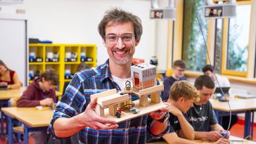 A man holds a wooden model in his hand, in the background a classroom in which pupils are trying out electronic components.