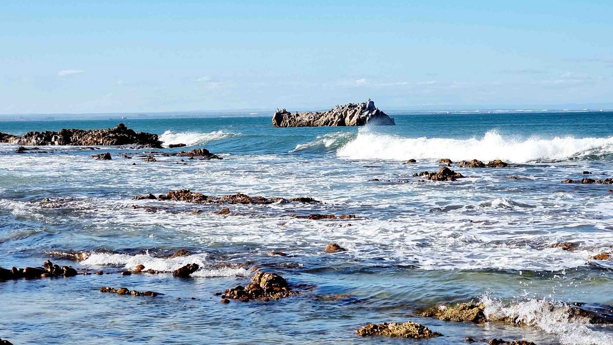 Eine Küstenlandschaft mit felsigen Abschnitten und Wellen. Im Hintergrund eine Insel oder ein Felsen.