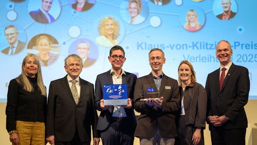 Six people, two women and four men, are standing in suits and blazers in front of a screen displaying the words “Klaus von Klitzing Prize”. The two men in the middle are holding two prizes in their hands. Everyone is smiling at the camera. 