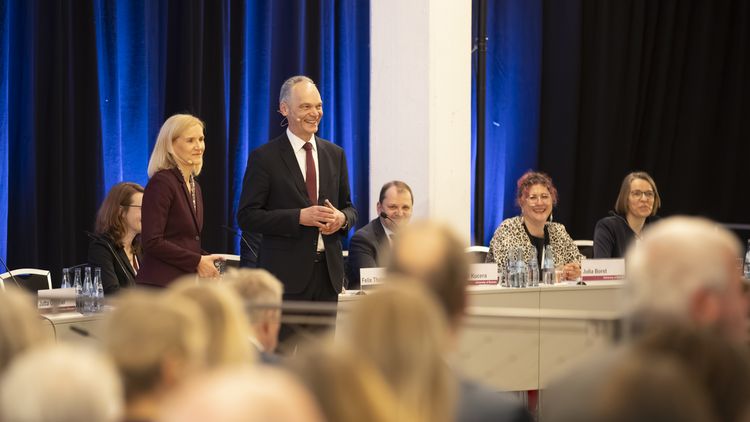 President Bruder and Vice-Chancellor Günther stand smiling in front of the audience, with a blue curtain in the background and other people seated at a table. 