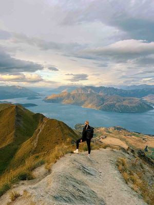 A woman stands on Roys Peak in New Zealand, overlooking a stunning mountain landscape with lakes and valleys. The sky is cloudy, but the sun is shining through.