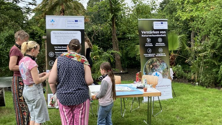 Auf einer Wiese im Botanischen Garten steht ein Tisch mit Infomaterial und zwei Aufstellern des Projekts. Mehrere Leute stehen vor dem Stand und schauen sich die Flyer an. 