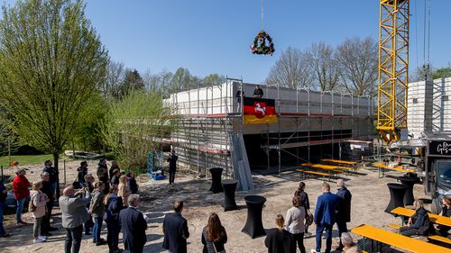 Long shot of the shell of the building with the topping-out wreath hanging above it. Some people are standing around the building.