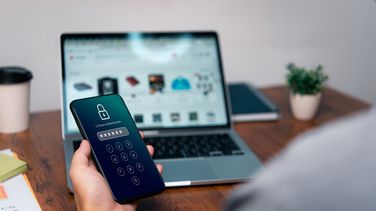 Im Hintergrund Laptop, auf dem undeutlich die Webseite eines Onlineshops zu sehen ist, im Vordergrund Hand, die ein Handy hält, auf dem ein verschlüsseltes Passwort eingegeben wurde. 