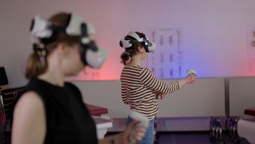 Two people with VR glasses over their eyes are standing in a seminar room and handling controllers.