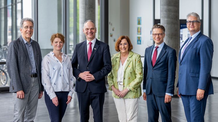 Two women and four men are standing in a row inside a building and smile into the camera. The men wear suits, one women wears a white blouse and dark-blue trousers, the other a green blazer and light-couloured trousers.