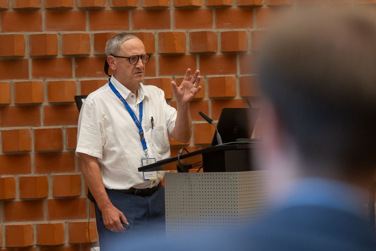 Ein Mann mit Brille und einem weißen Hemd steht hinter einem Pult und spricht. Er trägt ein Namensschild um den Hals. Er gestikuliert mit seinen Händen, während er spricht. Im Hintergrund ist eine rote Backsteinwand und vor ihm ein verschwommener Teil einer anderen Person zu sehen. Das Bild vermittelt den Eindruck einer Präsentation oder eines Vortrags während des Symposiums.