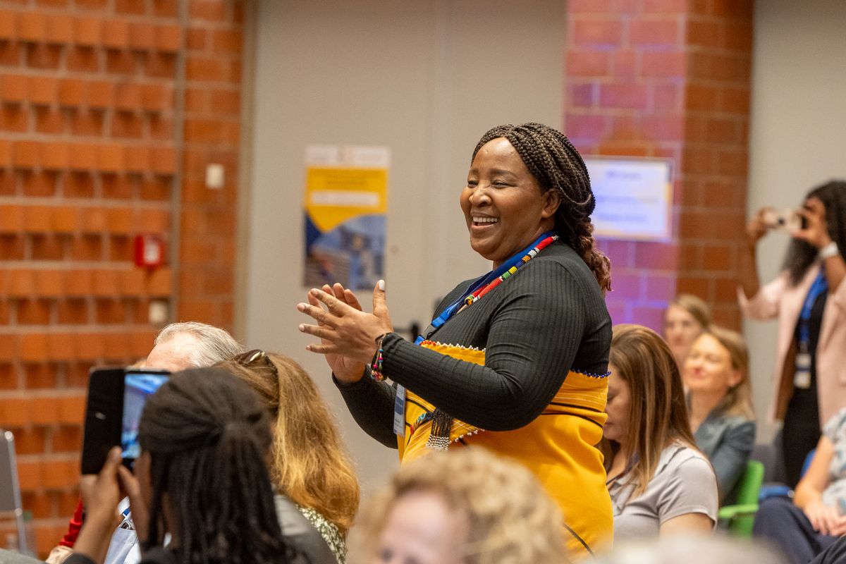 Das Bild zeigt eine Frau, die lächelnd und mit offenen Händen vor einer Gruppe von Menschen in die Hände klatscht. Sie trägt ein schwarzes Oberteil und einen gelben Rock mit farbenfrohen Mustern und eine bunte Halskette. Im Hintergrund sind weitere Personen zu sehen, einige von ihnen halten Smartphones hoch, möglicherweise um Fotos zu machen. Die Frau scheint sich in einer positiven und begeisterten Umgebung zu befinden,