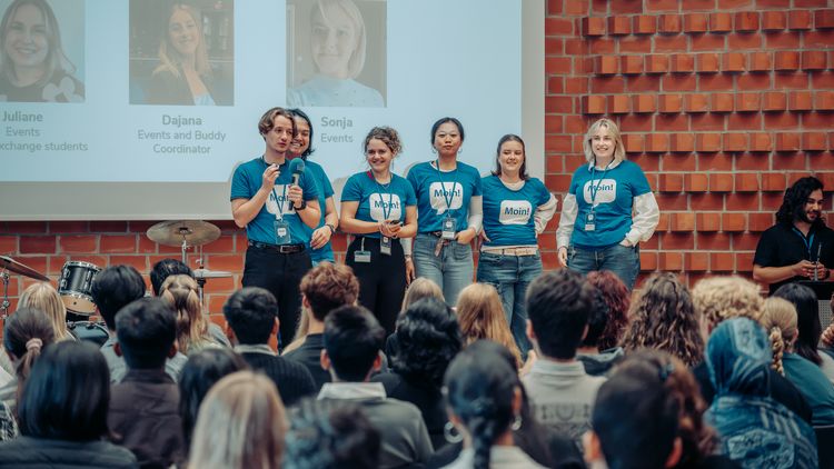 Sechs Tutoren für internationale Studierende in blauen „Moin!“-Shirts sprechen auf der Bühne vor dem Publikum und einer Präsentationsleinwand.