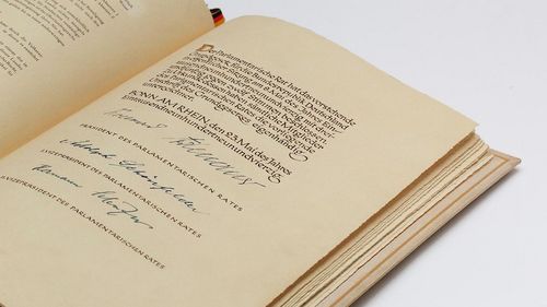 The picture shows an open book. It is the last page of the Basic Law with the three signatures. The bookmark is a ribbon in the colours black, red and gold.