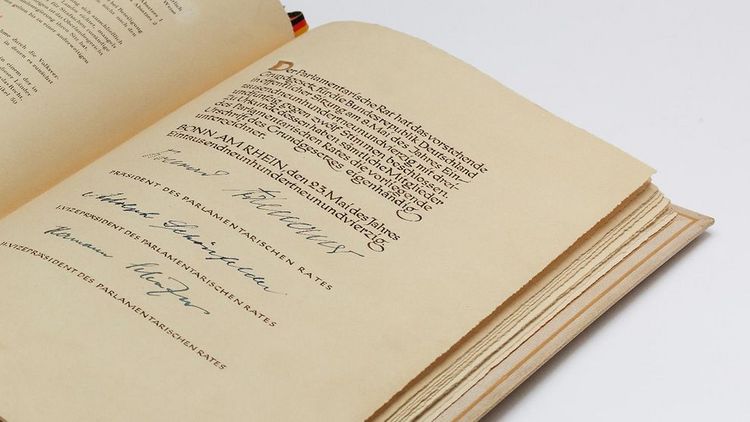 Das Bild zeigt ein aufgeklapptes Buch. Es ist die letzte Seite des Grundgesetzes mit den drei Unterschriften. Das Lesezeichen ist ein Bändchen in den Farben Schwarz-Rot-Gold.