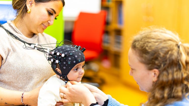 A young woman puts a cap with light-emitting diodes on a baby a few months old. The baby is sitting on its mother's lap.