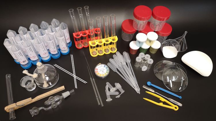Die Bestandteile des Experimentiersets sind auf dem Bild ausgebreitet: Reagenzgläser, Spatel, Klammern, ein Spiritusbrenner aus Glas, Chemikalien in röhrenförmigen Behältern, Becher mit Deckel etc. 