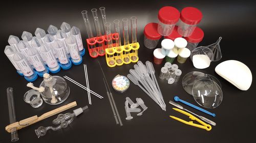 The components of the experimental set are spread out in the picture: Test tubes, spatulas, clamps, a glass spirit burner, chemicals in tubular containers, beakers with lids, etc.