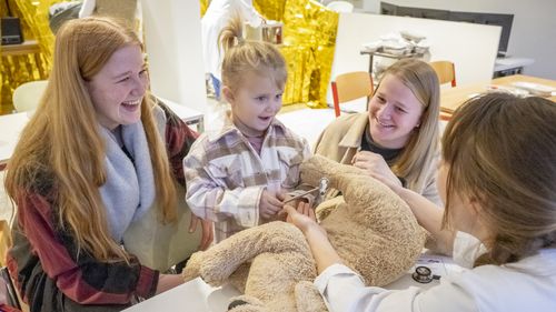 A little girl holds a reflex hammer. A large plush dog lies in front of her. Two smiling women sit to her right and left. Opposite her sits a woman in a smock.