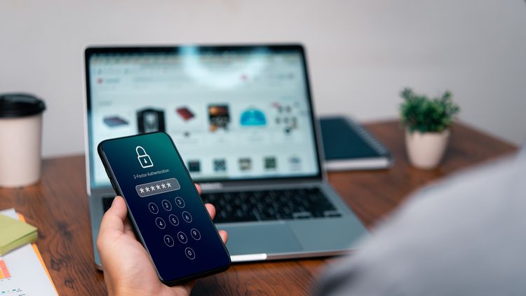 Im Hintergrund Laptop, auf dem undeutlich die Webseite eines Onlineshops zu sehen ist, im Vordergrund Hand, die ein Handy hält, auf dem ein verschlüsseltes Passwort eingegeben wurde. 