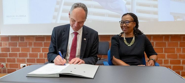 Das Bild zeigt zwei Personen, Vice-Chancellor Sibongile Muthwa (NMU) und Präsident Ralph Bruder (UOL), die an einem Tisch sitzen und ein Dokument unterzeichnen. Der Präsident der Universität Oldenburg schreibt mit einem Stift auf das Dokument. Die Vice-Chancellor der Nelson Mandela University lächelt und blickt auf das Dokument. Im Hintergrund ist ein Banner mit der Aufschrift "University of Oldenburg | President" und ein Bild des Universitätsgebäudes der NMU zu sehen. Das Bild dokumentiert die Unterzeichnung einer Vereinbarung im Rahmen des 25-jährigen Kooperationsjubiläums zwischen den beiden Universitäten.